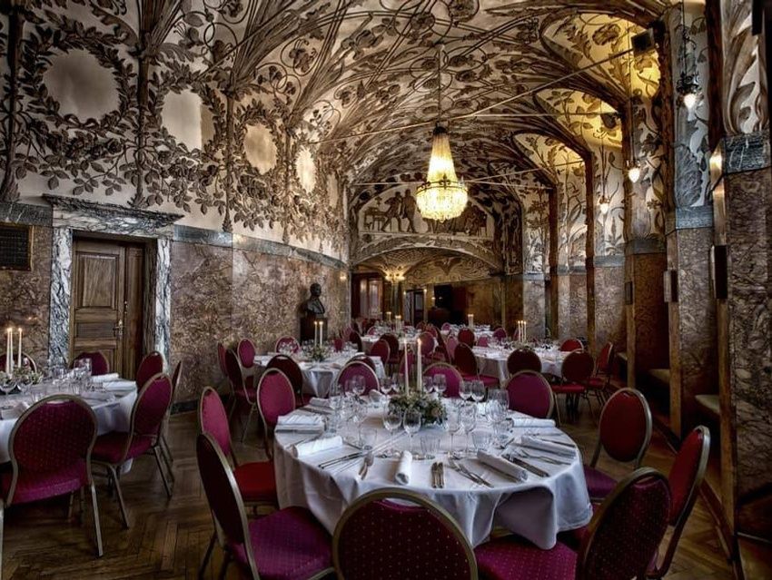 Elegant selskabslokale til et bryllup med hvælvet loft, detaljerede malerier, runde borde med hvide duge og røde stole, samt en stor lysekrone i midten.